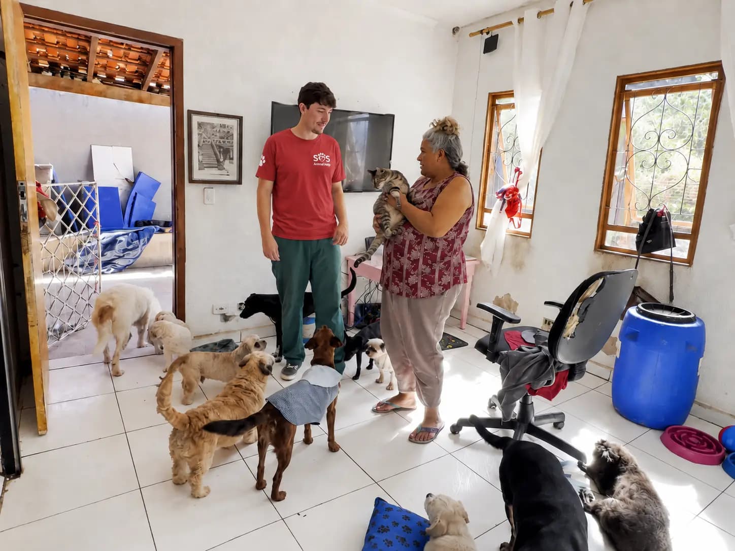 Protetor da SOS Animal Help, de camiseta vermelha, conversando com uma protetora que segura um gato no colo, dentro da casa onde ela acolhe dezenas de cães.